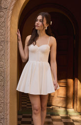 A woman is wearing a short, cream-colored dress with thin spaghetti straps and a fitted, textured bodice. The dress has a flared, pleated skirt that falls above her knees. She also has large, round earrings and a white hair accessory.
